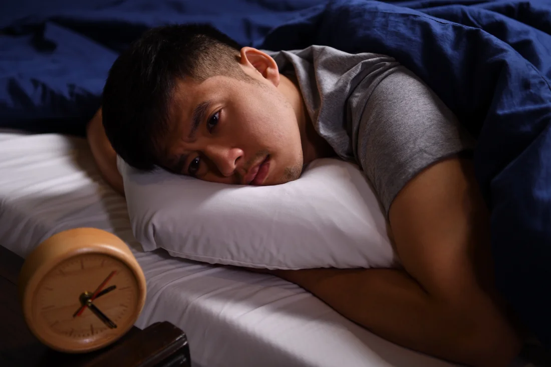 Man lying on his side in bed with a pillow under his head, looking tired while an orange alarm clock sits on the nightstand nearby.